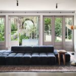 Modrá modulárna pohovka pred radom francúzskych okien pod svetlým stropom s čiernymi bodovými svietidlami.