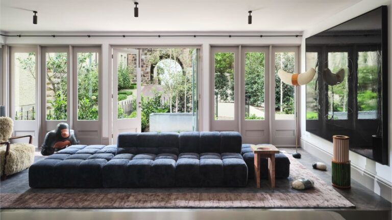 Modrá modulárna pohovka pred radom francúzskych okien pod svetlým stropom s čiernymi bodovými svietidlami.