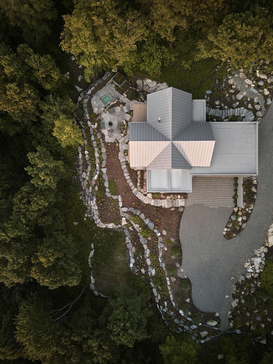 Pohľad zhora na modernú horskú chatu s plechovou strechou, kamennými terasami a prírodným lesným pozemkom.