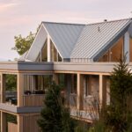 Priestranná exteriérová terasa s drevenými stĺpmi, skleneným zábradlím a tmavým záhradným nábytkom pod plochou strechou.