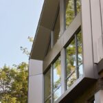 Fasáda moderného domu s vertikálnym sivým obkladom, lomenou líniou strechy a reflexnými sklenenými výplňami.