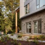 Prírodná kamenná terasa s bridlicovým chodníkom, nízkou vegetáciou a dreveným pódiom pri vstupe do domu.