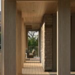 Krytá chodba s dreveným latkovým stropom, svetlou podlahou a robustnými nosnými piliermi zo svetlého dreva.