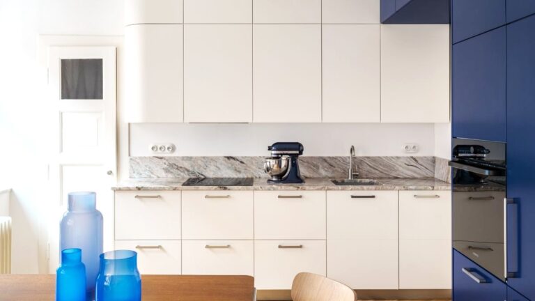 Biela minimalistická kuchyňa s kamennou pracovnou doskou, modrou vysokou skriňou a dubovými parketami.