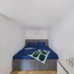 Sivá čalúnená posteľ s modrými vankúšmi umiestnená v bielom úzkom výklenku nad schodiskom.
