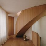 Chodba s drevenou podlahou, oblúkovitým schodiskom, bielou stenou, závesmi a hnedou kamennou plastikou.