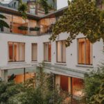 Budova s bielou fasádou, drevenými oknami, balkónmi s čiernym zábradlím a zeleným vnútroblokom.