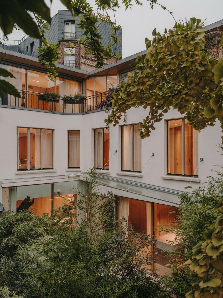 Budova s bielou fasádou, drevenými oknami, balkónmi s čiernym zábradlím a zeleným vnútroblokom.