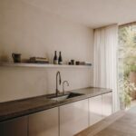 Kuchyňa s kamennou pracovnou doskou, nerezovými skrinkami, dizajnovou batériou, bielou policou a ľanovými závesmi.