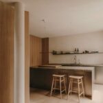 Kuchyňa s nerezovým ostrovom, kamennou doskou, drevenými stoličkami, bielym nosným stĺpom a lineárnym osvetlením.