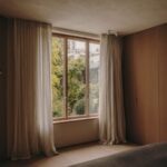 Spálňa so závesmi z ľanu, drevenými oknami s trojsklom, drevenou podlahou a výhľadom do zeleného vnútrobloku.