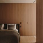 Spálňa s vstavanou skriňou z dubovej dyhy, čalúnenou posteľou s tmavosivou prikrývkou a čiernou lampou na čítanie.