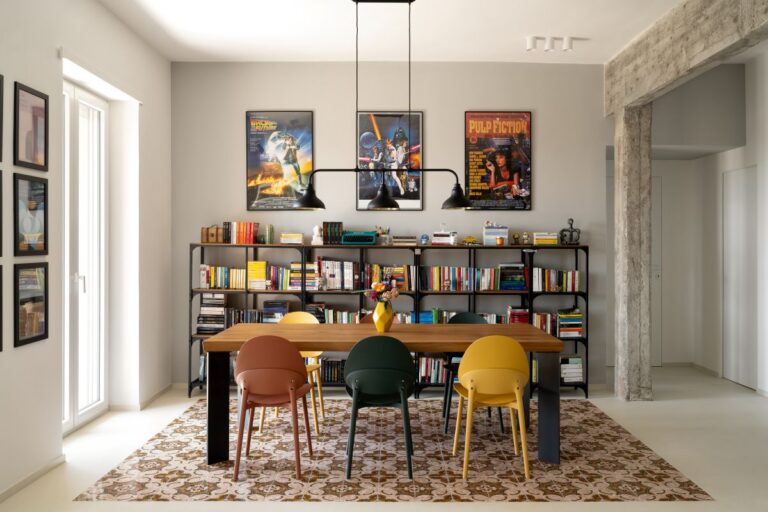 Filmové plagáty nad čiernou kovovou knižnicou za dreveným jedálenským stolom s farebnými dizajnovými stoličkami.