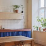 Kuchyňa s terakotovým kachličkovým ostrovom, modrým textilným závesom, bielou zástenou a svetlou drevenou skrinkou.