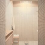 Sprchový kút s drobnou bielou mozaikou, priesvitný igelitový záves, drevená stolička a minimalistický úložný box.