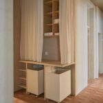 Chodba s drevenou policovou zostavou, svetlými textilnými závesmi, mobilnými bielymi kontajnermi a hnedou tehlovou podlahou.