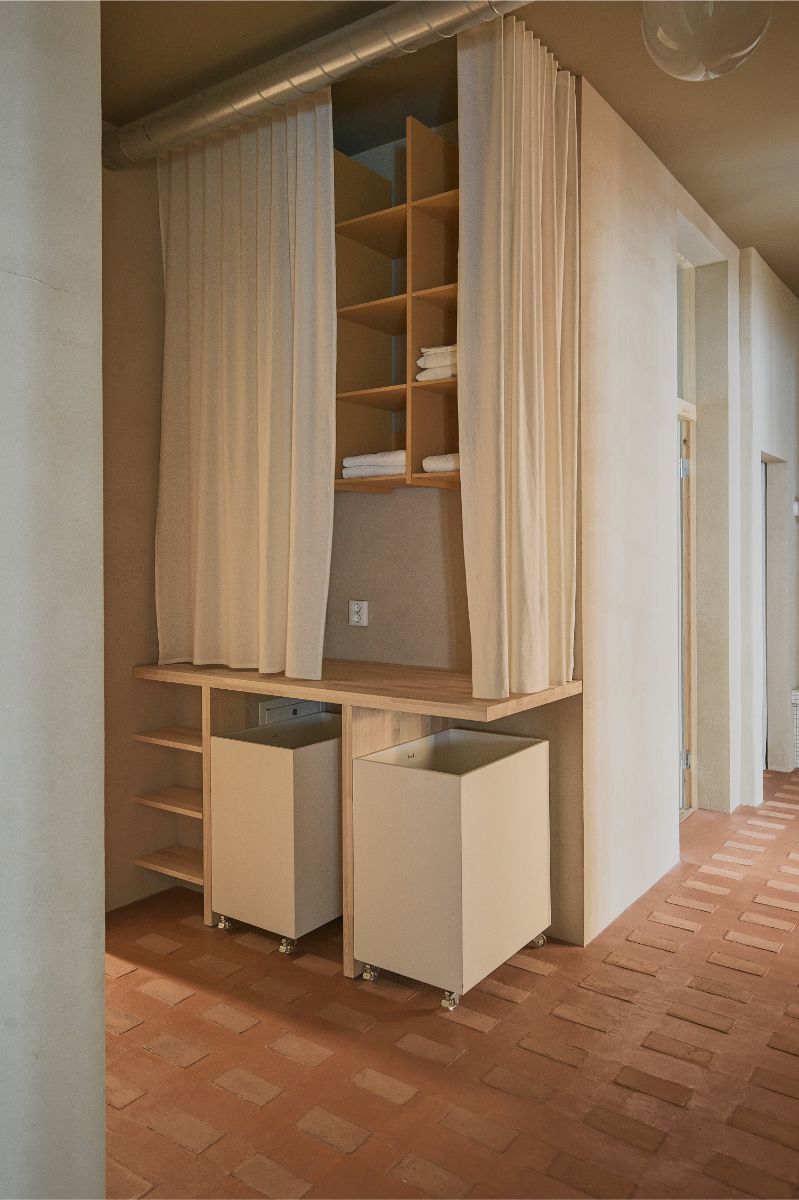 Chodba s drevenou policovou zostavou, svetlými textilnými závesmi, mobilnými bielymi kontajnermi a hnedou tehlovou podlahou.