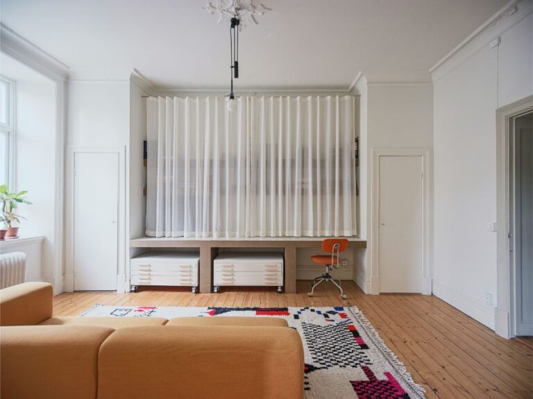 Pracovný kút v obývačke so stolom obloženým kachličkami a otvorenými policami, skrytými za bielymi záclonami.