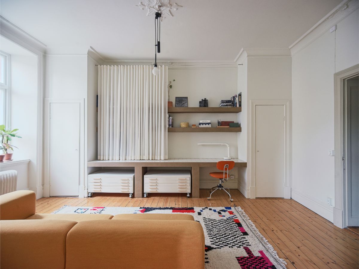 Pracovný kút v obývačke so stolom obloženým kachličkami a otvorenými policami, skrytými za bielymi záclonami.