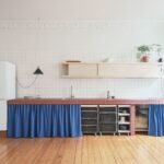 Kuchyňa s lineárnou linkou s tehlovým obkladom pracovnej dosky, policou s posuvnými dvierkami a modrým poodtiahnutým závesom a prepravkami na kolieskach.