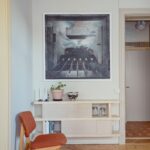 Obývacia izba s abstraktným obrazom, svetlým dreveným regálom, oranžovou retro stoličkou a zdobenou stropnou rímsou.