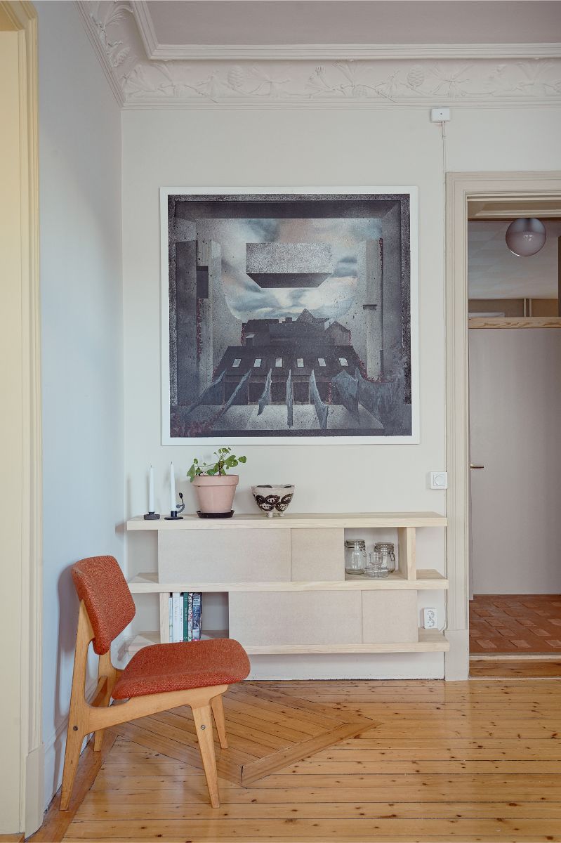 Obývacia izba s abstraktným obrazom, svetlým dreveným regálom, oranžovou retro stoličkou a zdobenou stropnou rímsou.