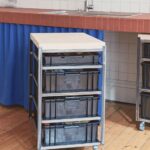 Kuchyňa s mobilným kovovým regálom, čiernymi plastovými zásuvkami, drevenou hornou doskou a lakovanou podlahou.