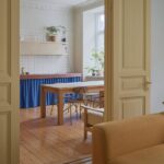 Pohľad cez krémové dvere do kuchyne s masívnym dreveným stolom a atypickou linkou s modrým závesom a obkladačkami na pracovnej doske.