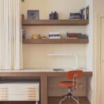 Pracovný kút so stolom s doskou s bielymi kachličkami, otvorenými policami, oranžovou kancelárskou stoličkou a krémovým nariaseným závesom.