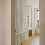 Dlhá biela kachličková polica s úložným priestorom skrytým za svetlými závesmi, drevenou podlahou a rastlinou v minimalistickej miestnosti.