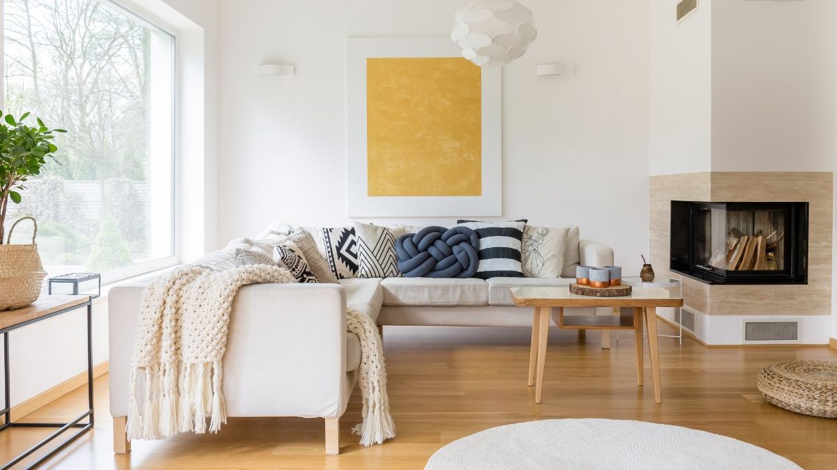 Svetlá rohová sedačka, žltý abstraktný obraz, drevený konferenčný stolík a biela tehlová obstavba krbu.