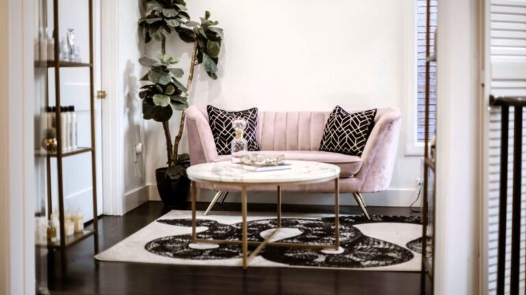 Ružová zamatová pohovka, čierne vzorované vankúše, mramorový stolík so zlatou podnožou a vysoký fikus.