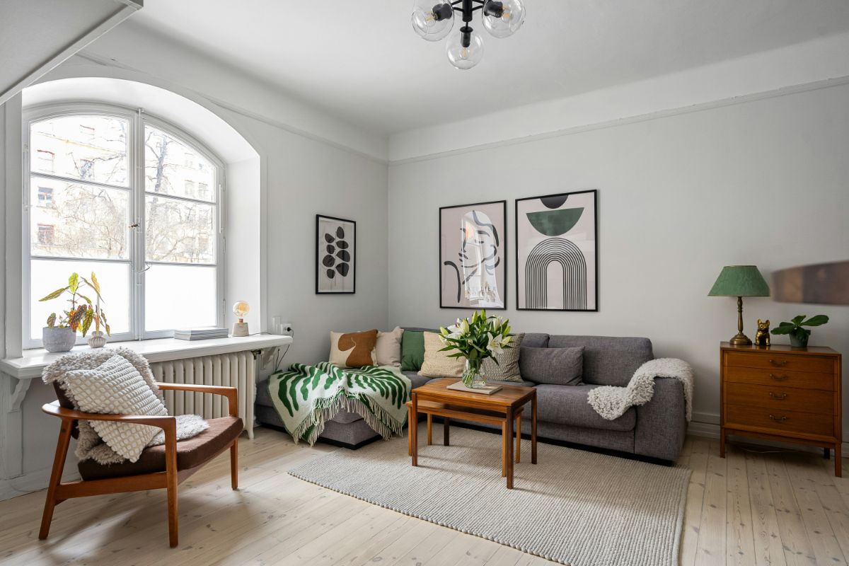 Sivá rohová pohovka, drevený retro nábytok, zelené interiérové doplnky a svetlá drevená podlaha.