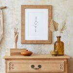 Drevená komoda, makramé dekorácia na stene, hnedý sklenený flakón a minimalistický čiernobiely obraz v boho štýle.