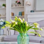 Biele tulipány v tyrkysovej sklenenej váze, sklenený konferenčný stolík a biela knižnica v pozadí.