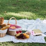 Svetlá bavlnená deka na tráve s prúteným piknikovým košom, ovocím a sklenenými pohármi v parku.