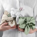 ri darčeky zabalené v bielej, béžovej a svetlozelenej bavlnenej látke s uzlom a ozdobnou zelenou halúzkou.