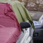 Zaparkované strieborné auto prekryté starou červenou a olivovozelenou látkou na ulici.