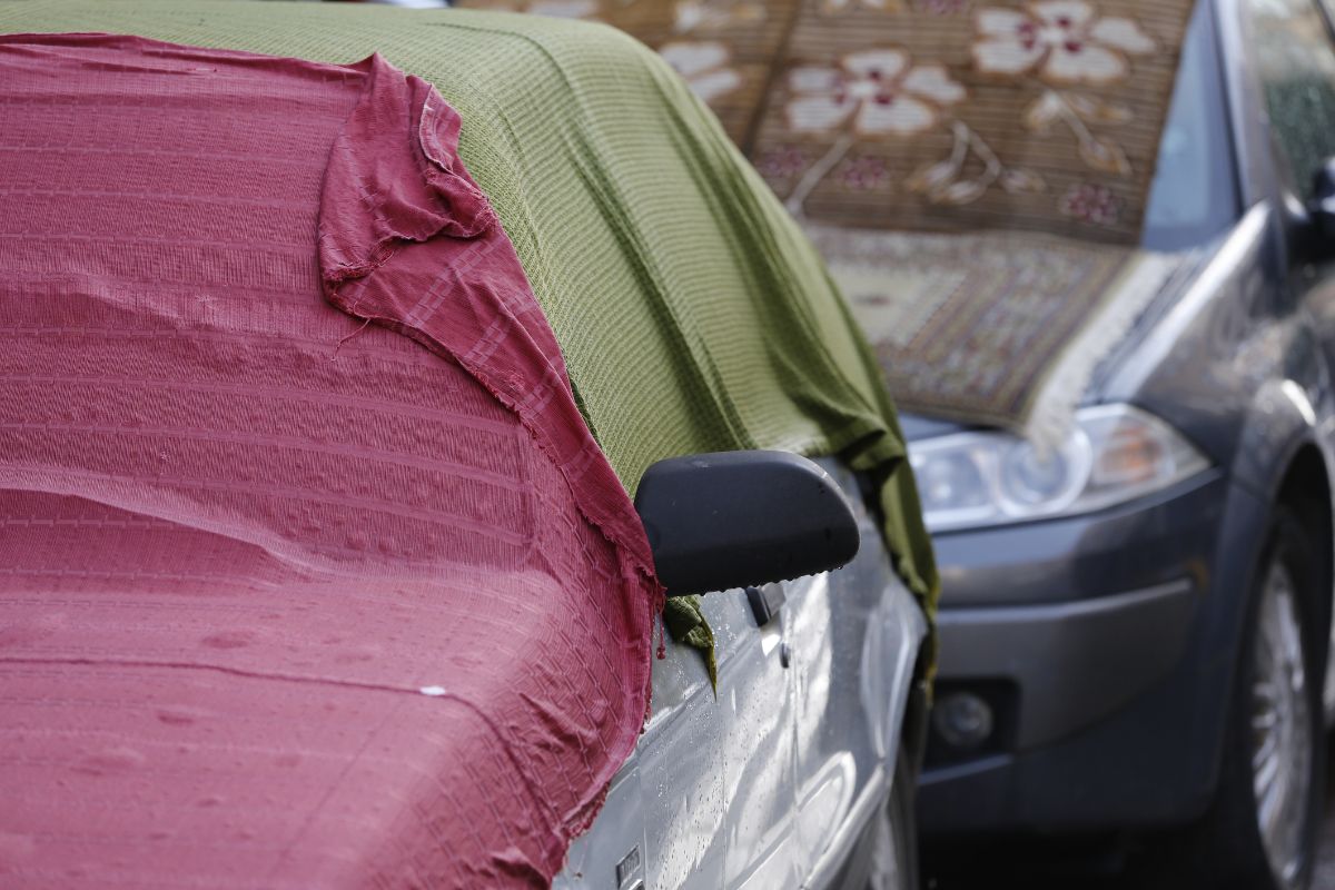 Zaparkované strieborné auto prekryté starou červenou a olivovozelenou látkou na ulici.