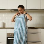 Žena v sivo-bielej pruhovanej kuchynskej zástere pred modernou bielou linkou s nerezovým drezom a batériou.