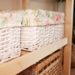 Biele prútené košíky vystlané svetlou látkou s ružovým kvetinovým vzorom na drevenej polici.
