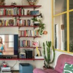 Obývačka s červenou otvorenou knižnicou, staroružovou čalúnenou pohovkou, vzorovanou dlažbou a žltým kovovým okenným rámom.