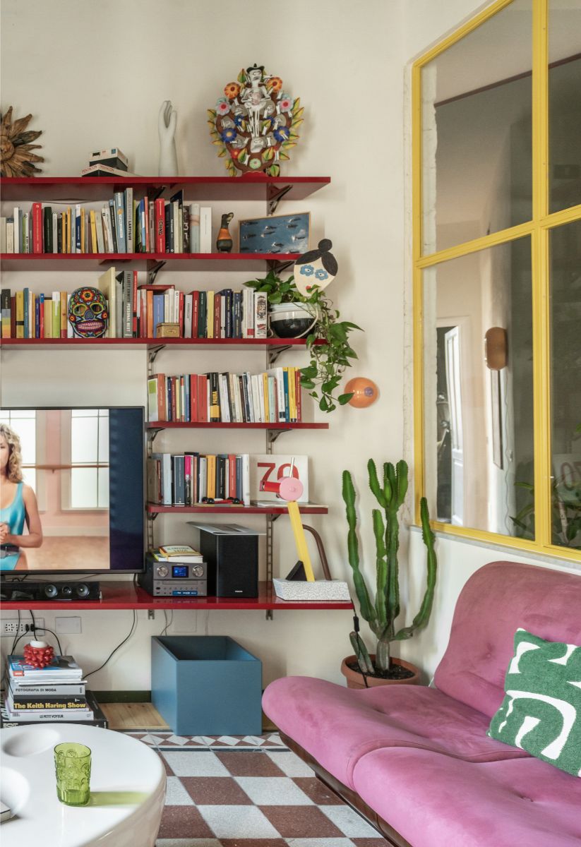 Obývačka s červenou otvorenou knižnicou, staroružovou čalúnenou pohovkou, vzorovanou dlažbou a žltým kovovým okenným rámom.