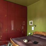 Spálňa s tmavočervenou lakovanou skriňou, olivovozelenými stenami, drevenou retro posteľou a dizajnovým stropným ventilátorom.