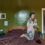 Žena na drevenej vintage posteli s hnedou bielizňou pred sýtozelenou stenou a bielymi kazetovými dverami v spálni.