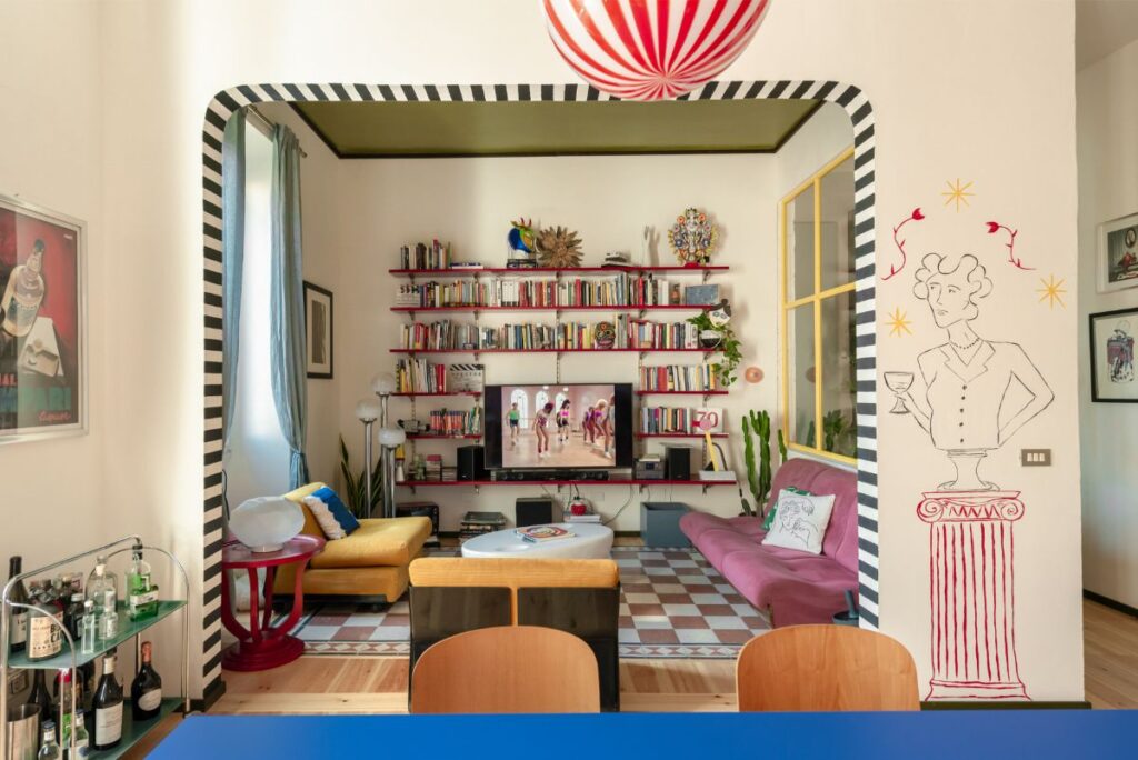 Farebný interiér s červenými policami, žltým kreslom, ružovou pohovkou a čiernobielym pruhovaným lemovaním výrazného prekladu.