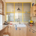 Svetlá drevená kuchynská linka, mramorová pracovná doska, biele keramické dvojumývadlo a žlté kovové okná s rastrom.