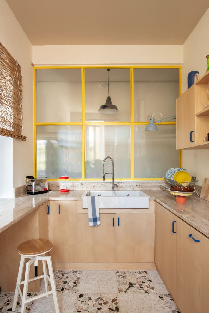 Svetlá drevená kuchynská linka, mramorová pracovná doska, biele keramické dvojumývadlo a žlté kovové okná s rastrom.