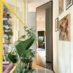 Drevená palubová podlaha, vysoké izbové rastliny, biele steny s obrazmi a veľké otočné zrkadlo v svetlej chodbe.