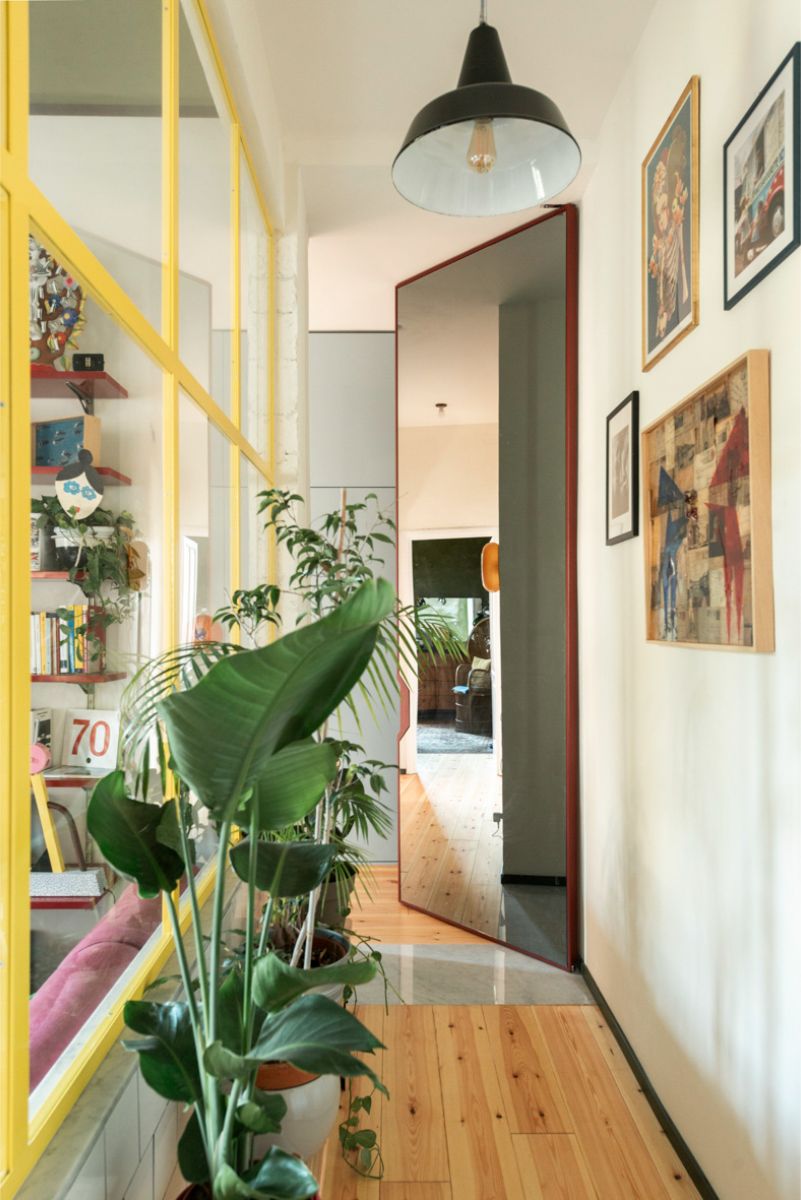 Drevená palubová podlaha, vysoké izbové rastliny, biele steny s obrazmi a veľké otočné zrkadlo v svetlej chodbe.